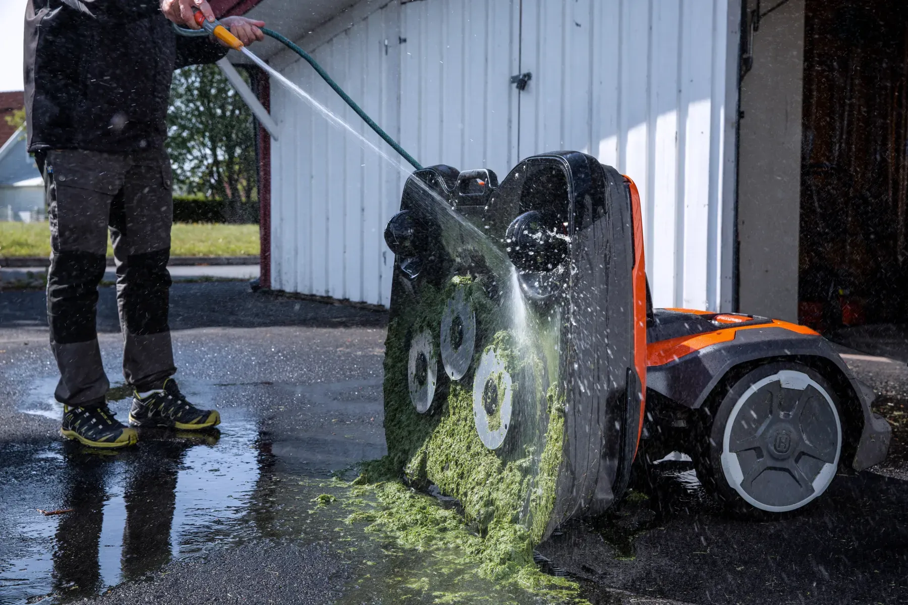 En person sprutar vatten med en slang för att rensa bort gräsklipp från undersidan av en orange robotgräsklippare på en uppfart utanför ett vitt garage.