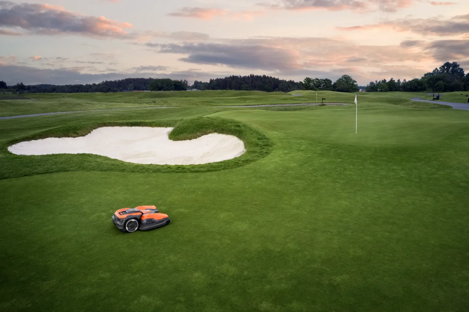 En robotgräsklippare trimmar gräset nära en sandbunker på en golfbana i solnedgången, med en flagga som markerar hålet i fjärran och träd i horisonten.