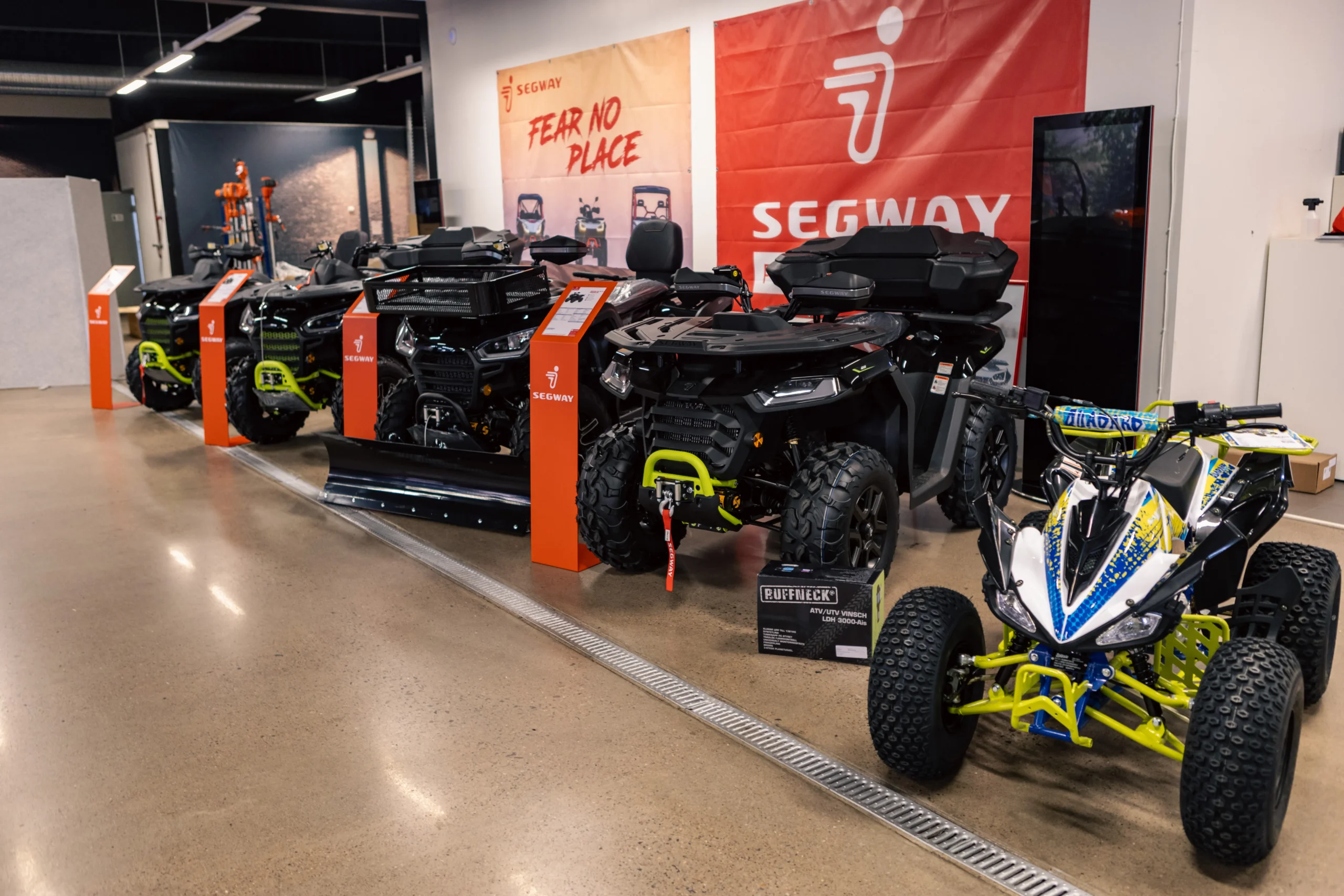 R6II9936 I en utställningslokal står flera svarta terrängfordon av märket Segway på rad, med orange Segway-skyltar och -banderoller bakom, och en mindre vit och blå fyrhjuling till höger.