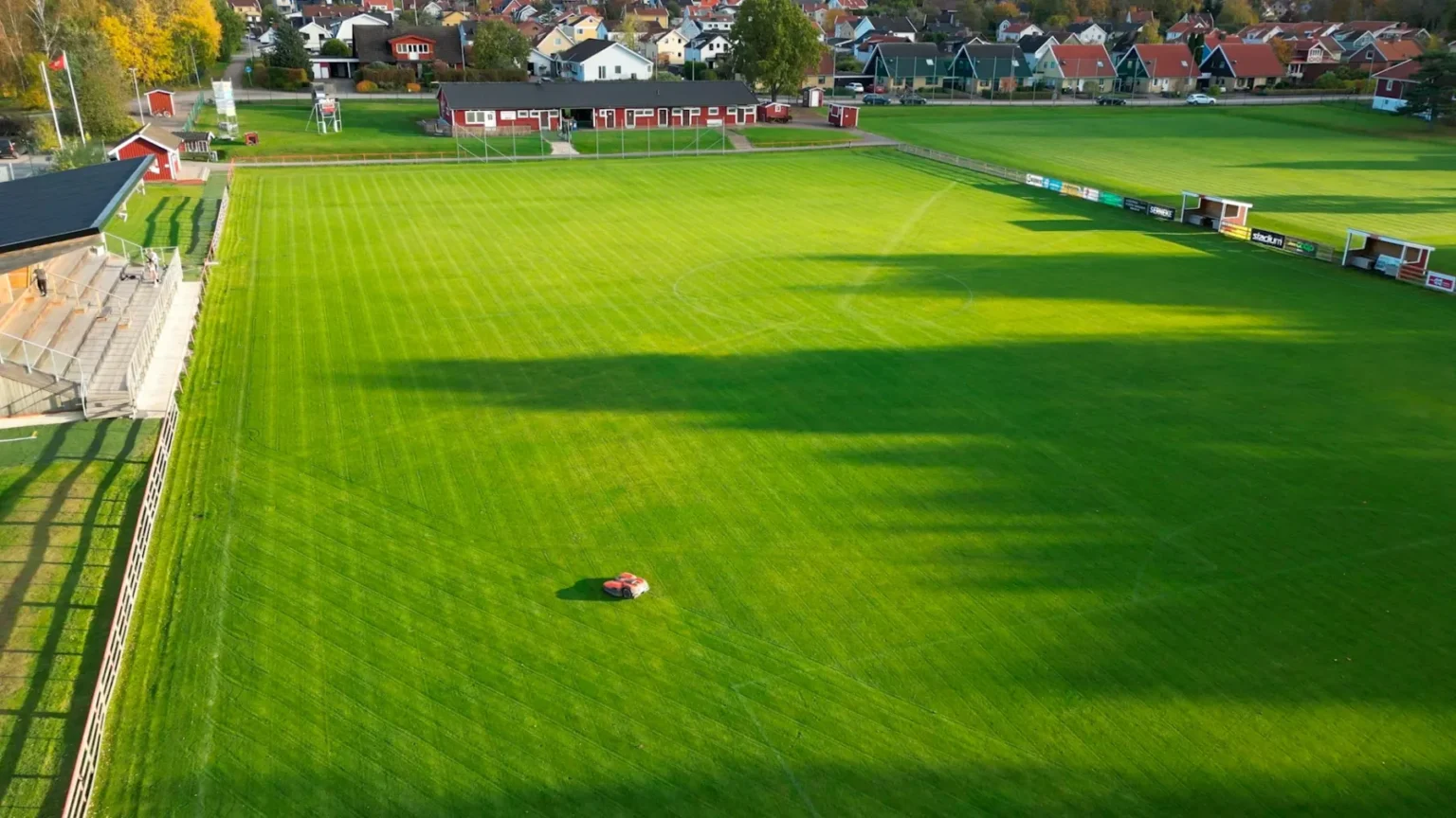 Flygfoto över en välskött fotbollsplan med ljusgrönt gräs, omgiven av ett staket och läktare, i ett bostadsområde med hus och träd. En liten röd gräsklippare syns på planen.