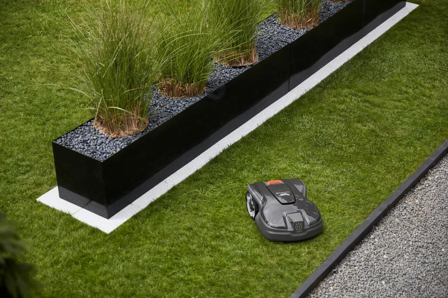En robotgräsklippare klipper gräs nära en svart rektangulär plantering fylld med dekorativa gräs och svarta stenar, kantad av en vit remsa och med grus synligt på sidan.