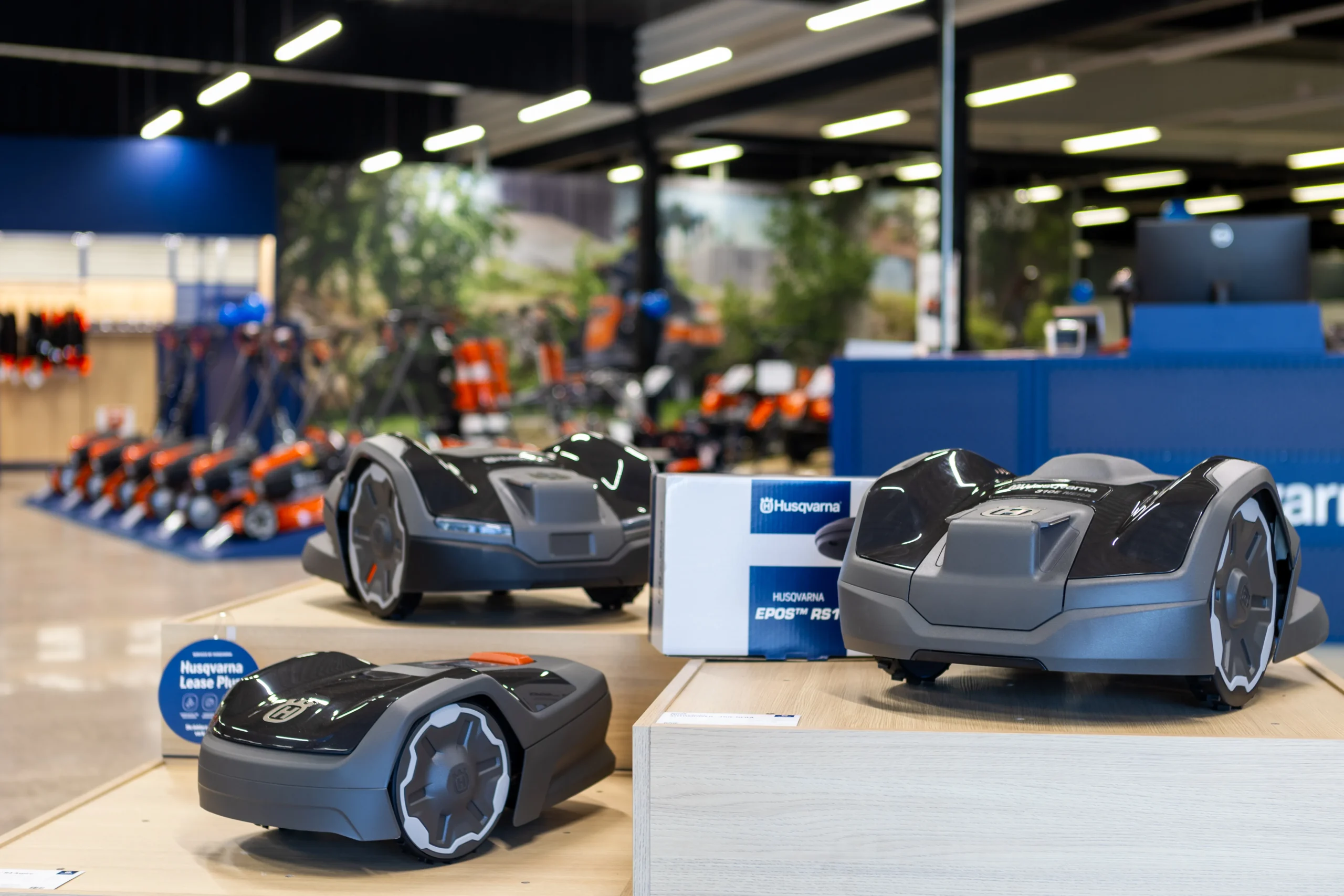 Tre robotgräsklippare visas på trästativ i ett väl upplyst utställningsrum, med Husqvarnas varumärke synligt och annan trädgårdsutrustning i bakgrunden.