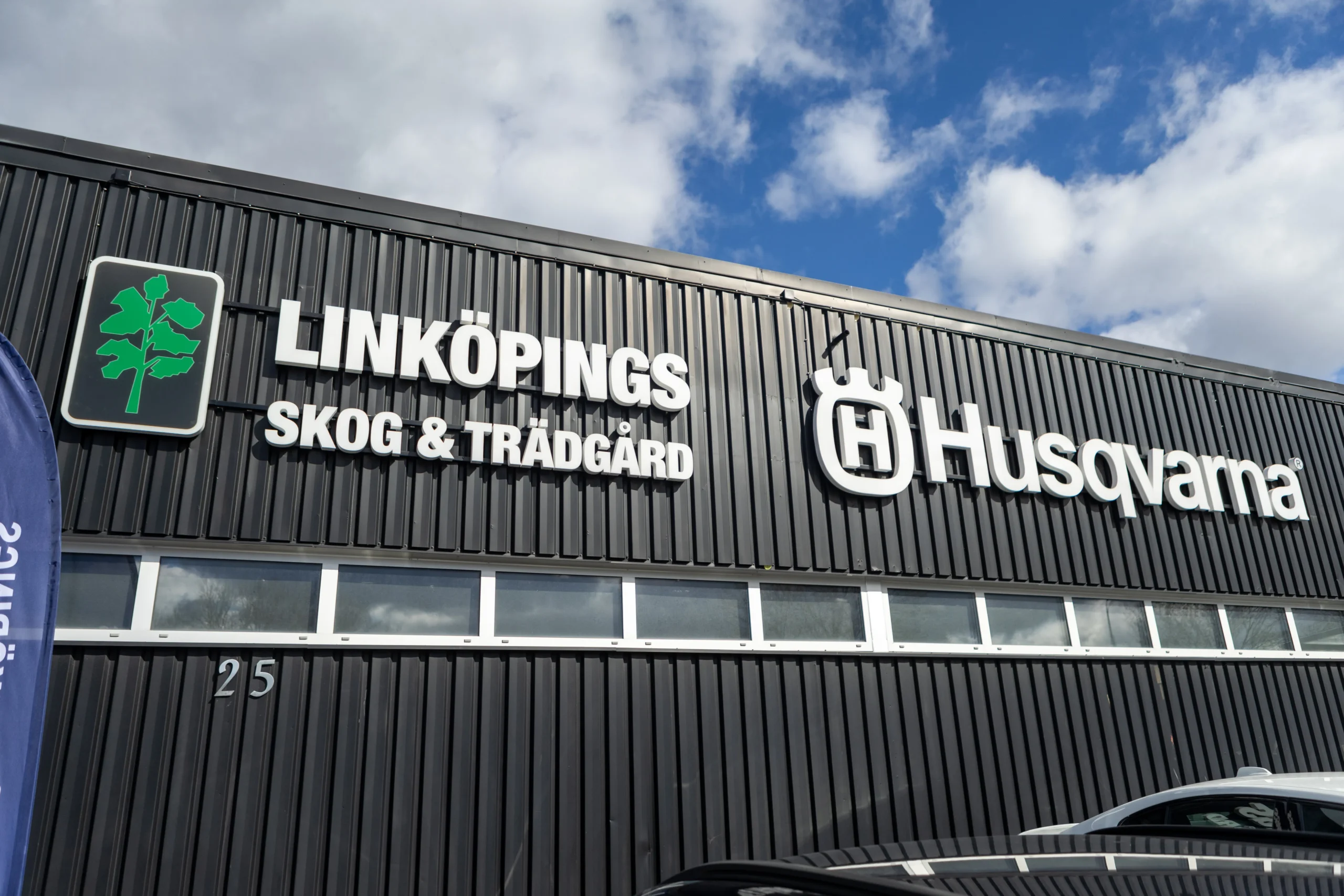 En svart byggnadsfasad med skyltarna "LINKÖPINGS SKOG & TRÄDGÅRD" och "Husqvarna" i vita bokstäver, tillsammans med deras logotyper. Himlen med moln syns ovanför byggnaden.