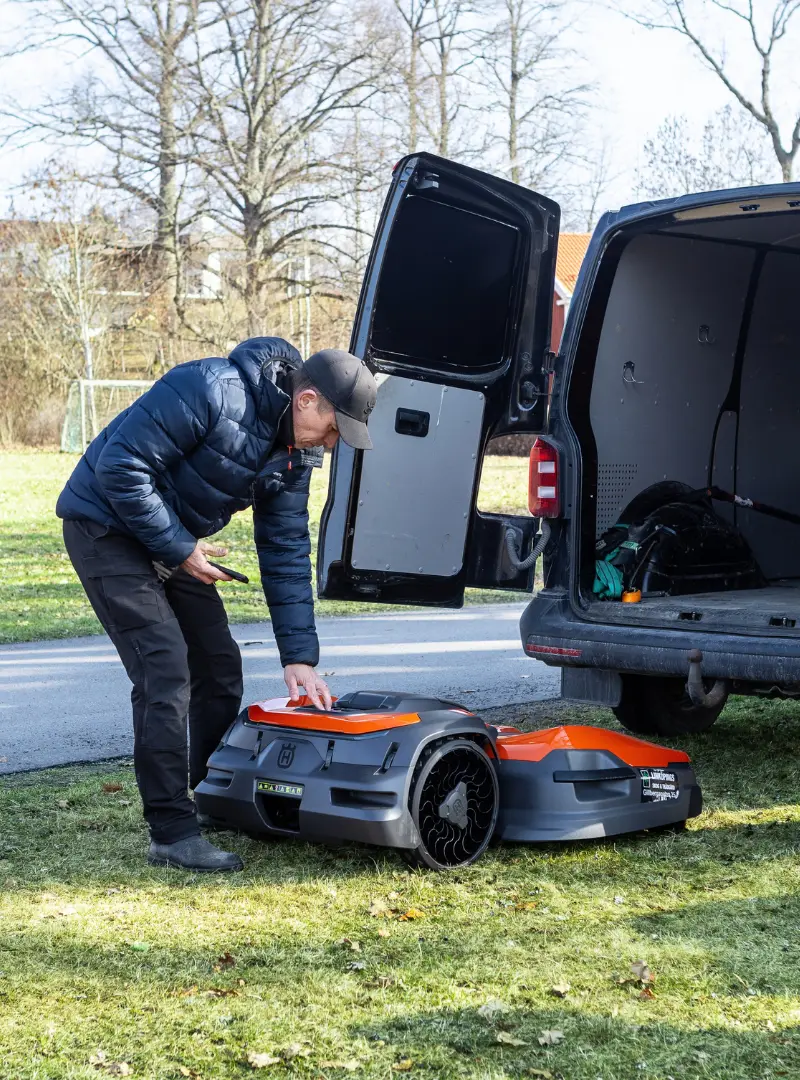 En person i svart jacka och keps tittar på en robotgräsklippare bredvid en öppen skåpbil som står parkerad på gräset, med träd och ett fotbollsmål i bakgrunden.