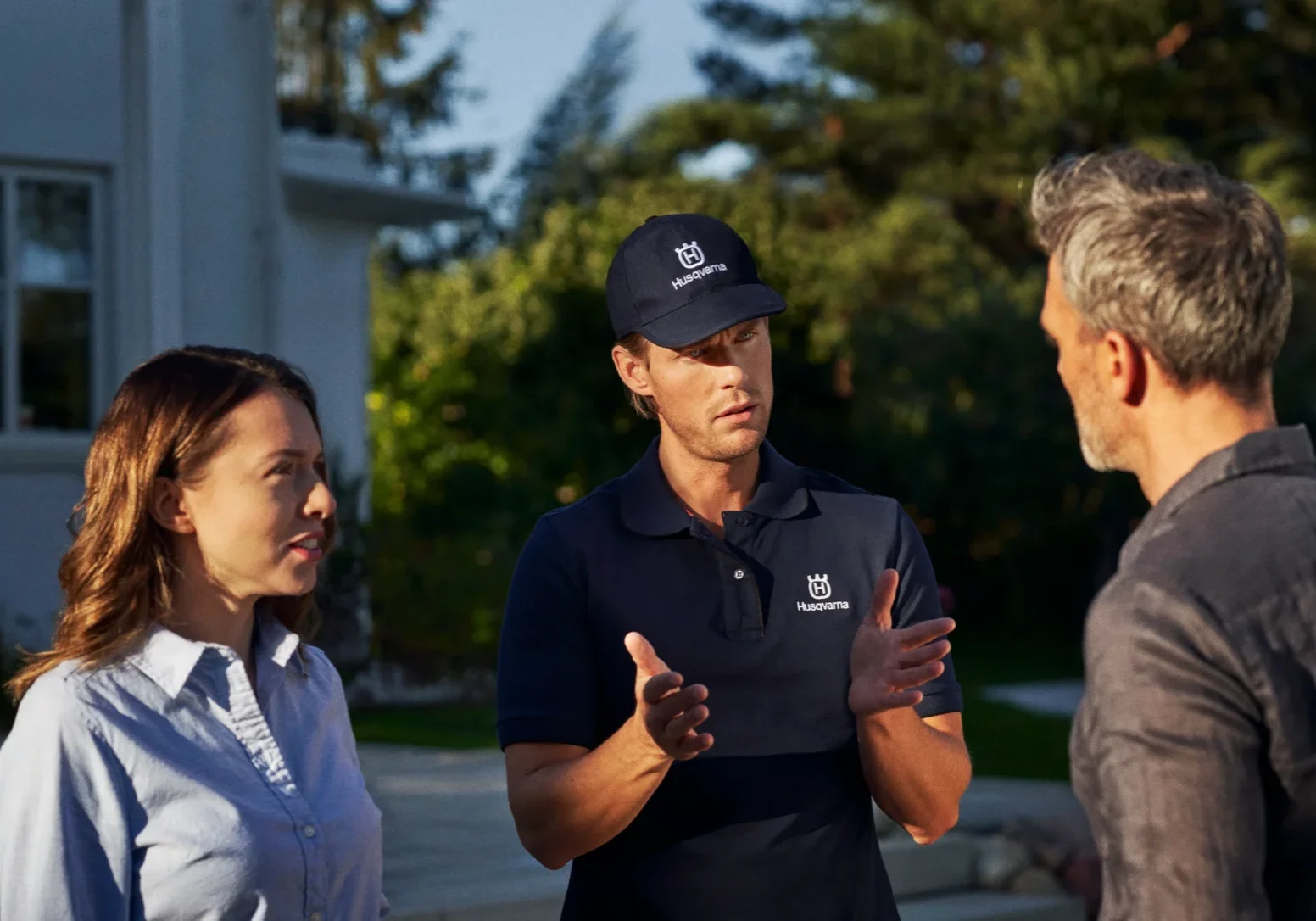 En kvinna och en man står utanför ett hus och pratar med en budbärare i marinuniform och keps. Arbetaren gestikulerar med händerna medan han förklarar något. Träd och en del av huset syns i bakgrunden.